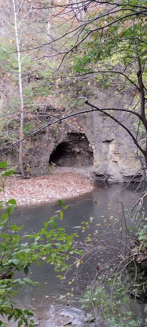 A serene river scene with a rocky cave entrance surrounded by trees and autumn leaves.