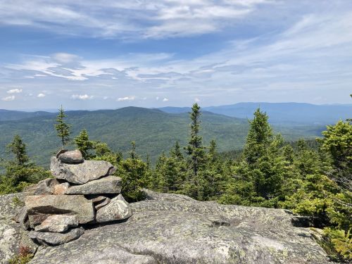 A rocky outcrop with a stacked stone cairn overlooks a lush green valley and distant mountains under a blue sky.