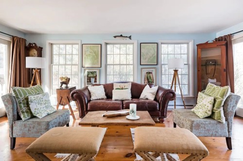 Cozy living room with a brown leather sofa, patterned chairs, wooden coffee table, and large windows letting in natural light.