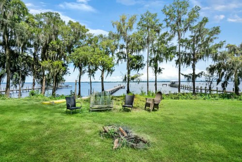 A serene lakeside view with chairs arranged around a fire pit, surrounded by trees and a dock in the background.