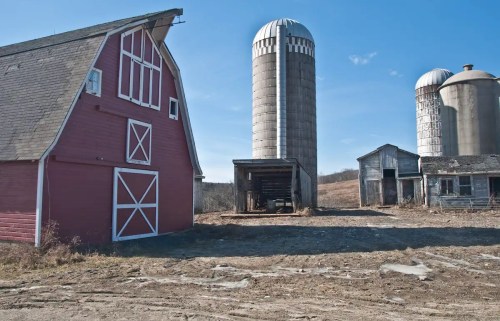 A red barn and silos stand in a rural landscape under a clear blue sky, with a weathered building nearby.