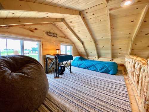 Cozy attic space with wooden walls, a blue sleeping bag, bean bag chair, and a small table by the windows.