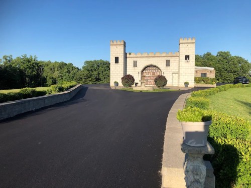 A castle-like entrance with a stone facade and a winding driveway, surrounded by greenery under a clear blue sky.