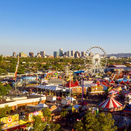 Aerial view of a vibrant fairground with rides, games, and a skyline in the background under a clear blue sky.