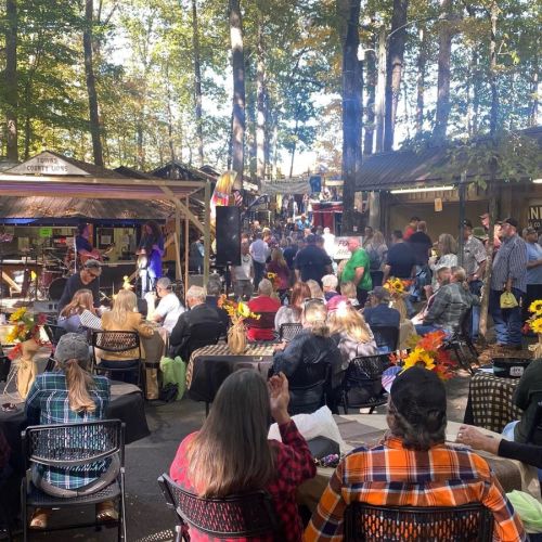 A crowd gathers at an outdoor event in a wooded area, with tables decorated with sunflowers and a stage in the background.