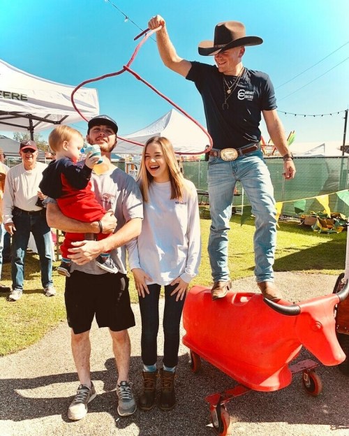 A cowboy on a red toy bull entertains a family at a fair, with a child in the father's arms and smiles all around.
