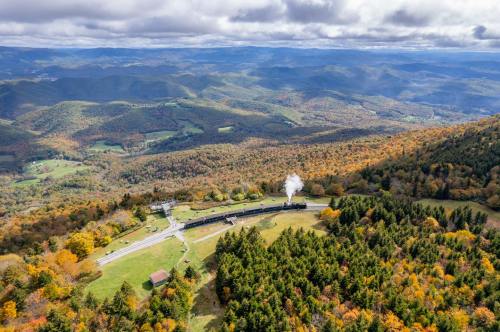 Aerial view of a steam train in a valley surrounded by colorful autumn foliage and rolling mountains under a cloudy sky.