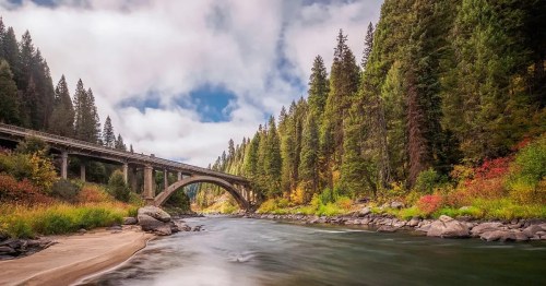 A serene river flows under a bridge, surrounded by lush green trees and colorful autumn foliage against a cloudy sky.