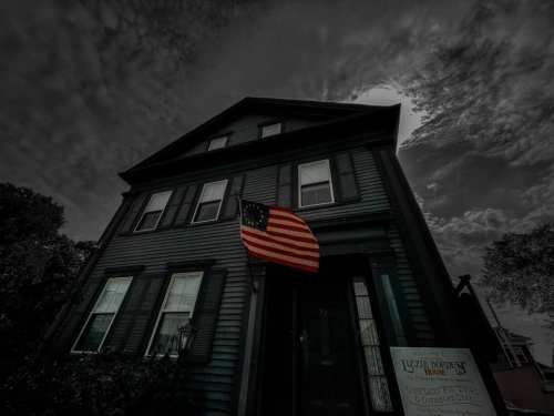 A dark, moody image of a historic house with an American flag hanging outside under a cloudy sky.