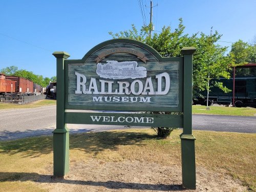 Sign for a Railroad Museum, featuring the word "WELCOME" and surrounded by greenery and railroad cars in the background.