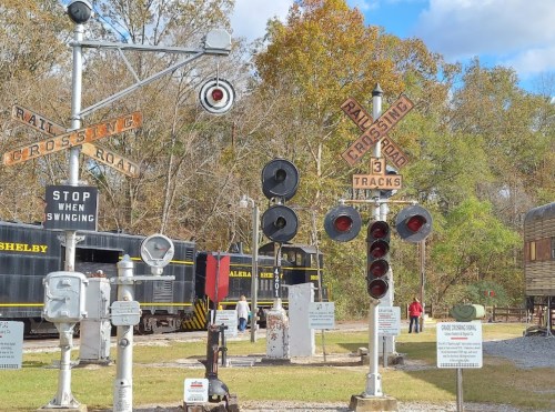 Railroad crossing signals and tracks in a museum setting, with vintage trains and trees in the background.