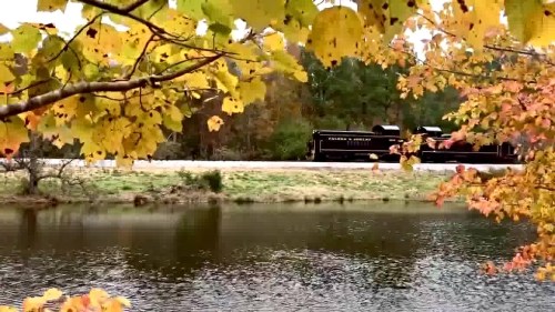 A vintage train travels alongside a serene lake, framed by vibrant autumn leaves.