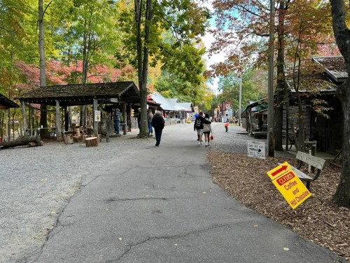 A scenic pathway in a wooded area with shops, people walking, and colorful autumn leaves. A sign for coffee is visible.