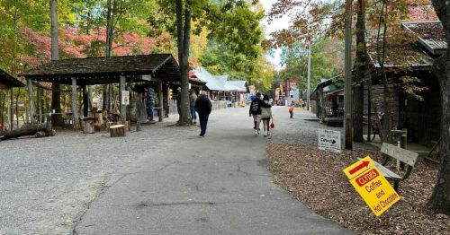 A scenic pathway in a wooded area with shops, people walking, and colorful autumn leaves. A sign for coffee is visible.