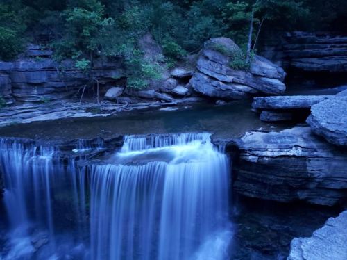 A serene waterfall cascades over rocky ledges, surrounded by lush greenery in a tranquil, dimly lit setting.
