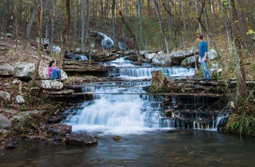 A serene waterfall cascades over rocks, with two people enjoying the peaceful nature scene in a forested area.