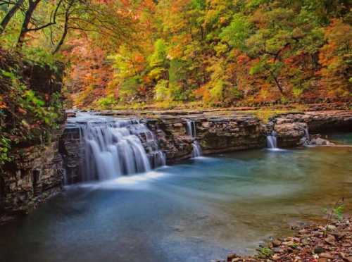 A serene waterfall cascades over rocky ledges, surrounded by vibrant autumn foliage and a calm, clear pool.