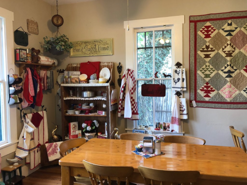 Cozy kitchen with a wooden table, quilted wall decor, and shelves filled with dishes and vintage kitchen items.