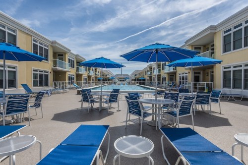 A sunny pool area with blue umbrellas, lounge chairs, and tables, surrounded by yellow buildings.