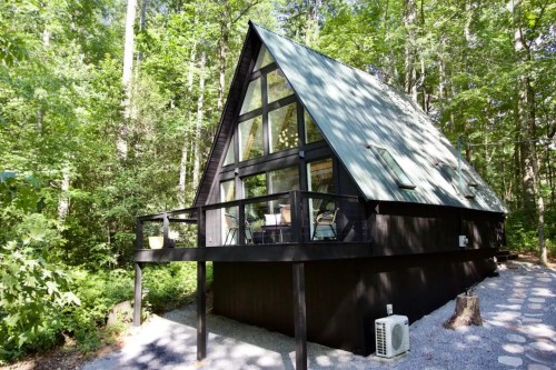 A modern A-frame cabin surrounded by lush green trees, featuring a large deck and large windows.