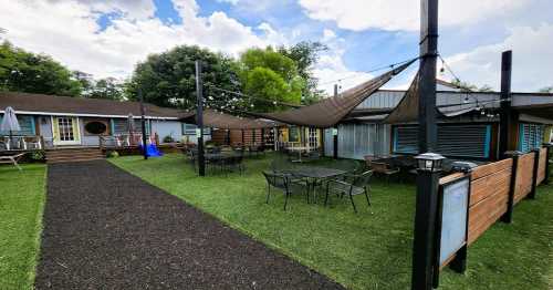 Outdoor seating area with tables, chairs, and shade sails, surrounded by greenery and a building in the background.