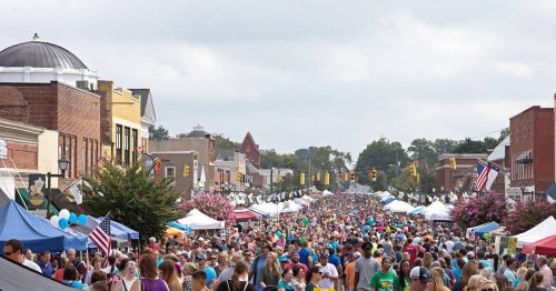 A bustling street fair with large crowds, colorful tents, and festive decorations under a cloudy sky.