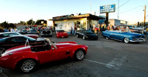 A lively drive-in scene with classic cars parked outside a diner, people mingling, and a clear blue sky.