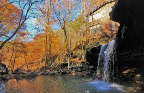 A serene waterfall cascades into a pond, surrounded by autumn foliage and a rustic cabin in the background.