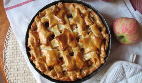 A freshly baked apple pie with a star-shaped lattice crust, placed on a white cloth next to a whole apple.