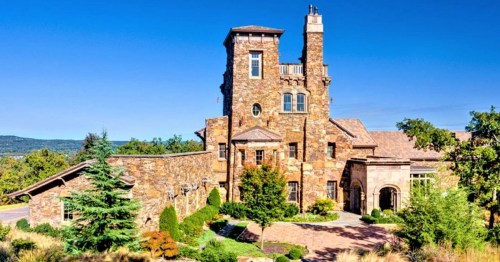 A large stone mansion with a tower, surrounded by lush greenery and a clear blue sky.