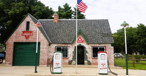 Historic Conoco gas station with a brick building, green accents, and vintage gas pumps, set against a tree-lined background.