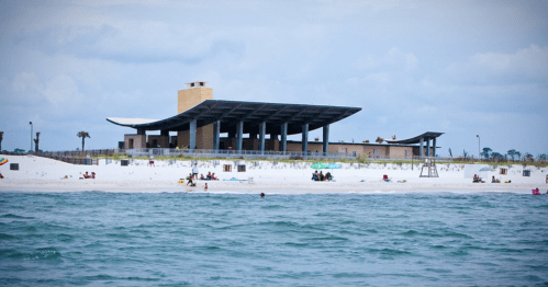 A beach scene featuring a modern building with a large roof, surrounded by white sand and people enjoying the water.