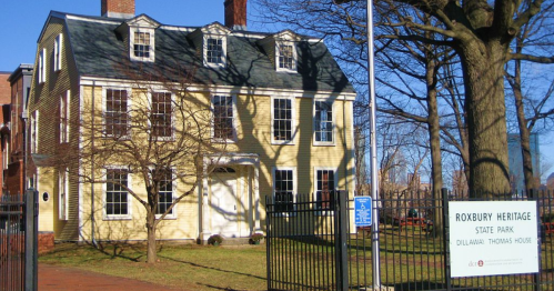 Historic yellow house with multiple windows, surrounded by a fence, next to a sign for Roxbury Heritage State Park.