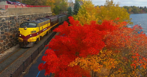 A vintage train travels along a scenic route with vibrant autumn foliage in red, orange, and yellow.