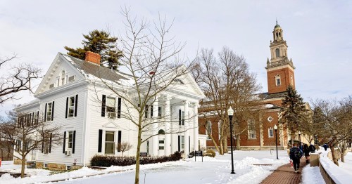 A snowy campus scene featuring a white historic building and a clock tower in the background, with students walking by.