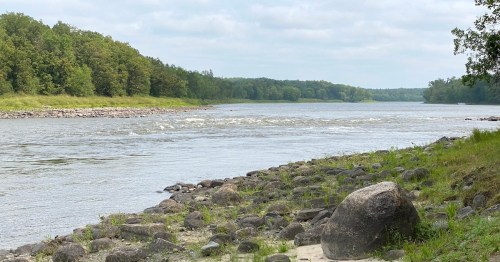 A calm river flows through a lush green landscape, with rocky banks and trees lining the shore under a cloudy sky.