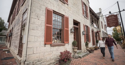A couple walks hand in hand past a historic stone building with red shutters and a sign for a restaurant and gift shop.