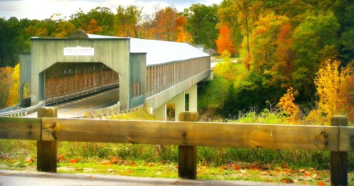 A scenic view of a covered bridge surrounded by vibrant autumn foliage and a wooden fence in the foreground.