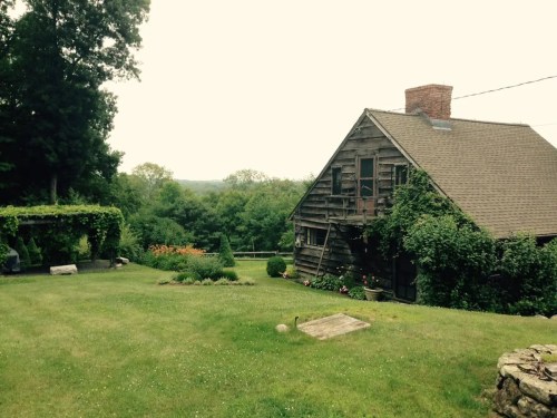 A rustic wooden house surrounded by greenery and a well-maintained lawn, with distant hills in the background.