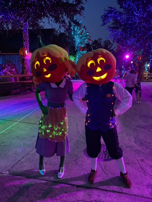 Two characters with pumpkin heads and festive outfits pose in a colorful, illuminated setting at night.