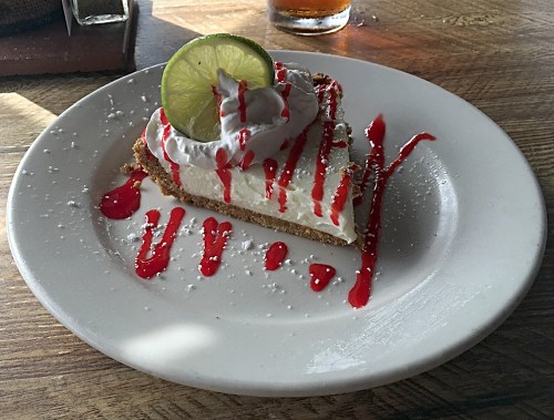 A slice of key lime pie topped with whipped cream and a lime wedge, drizzled with raspberry sauce on a white plate.