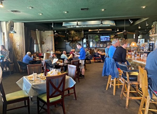 A busy restaurant interior with diners at tables and a bar, featuring rustic decor and a TV in the background.