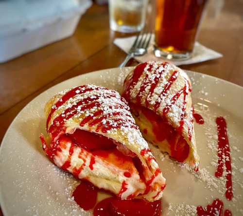 Two dessert crepes filled with cream and strawberries, drizzled with red sauce and dusted with powdered sugar on a plate.