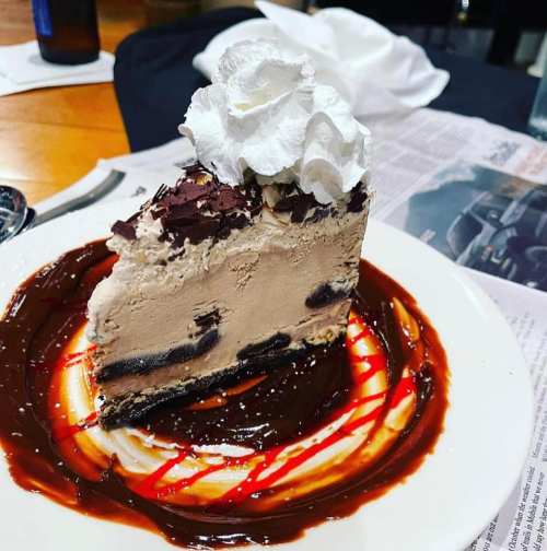 A slice of chocolate mousse cake topped with whipped cream, drizzled with chocolate and raspberry sauce on a plate.