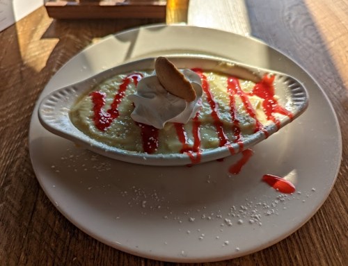 A dessert in a white dish topped with whipped cream, a cookie, and drizzled with red sauce on a wooden table.