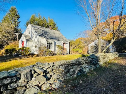 A quaint white house surrounded by trees and a stone wall, set against a clear blue sky.