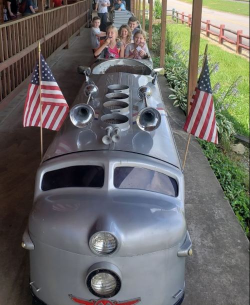 A vintage silver toy train with American flags, parked beside children smiling and posing for a photo.