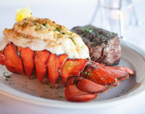 A plate featuring a lobster tail and a cooked steak, garnished with herbs and a lemon wedge.