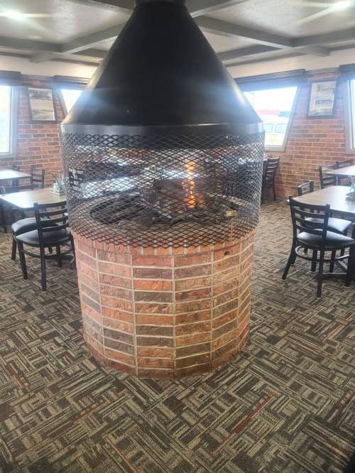 A cozy restaurant interior featuring a central brick fireplace with a metal grate, surrounded by tables and chairs.
