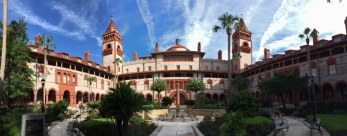 Historic building with red brick architecture, palm trees, and a central courtyard under a blue sky with clouds.